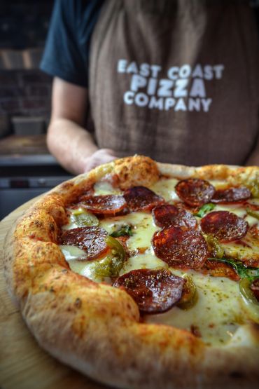 Close-up of a wood-fired pepperoni pizza held by a team member in a branded apron