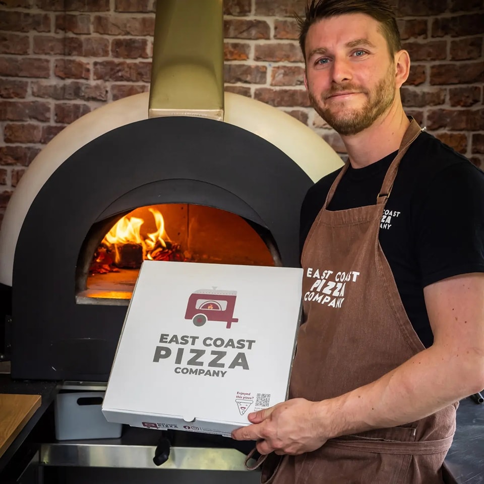 Chris holding an East Coast Pizza Company pizza box beside the wood-fired oven