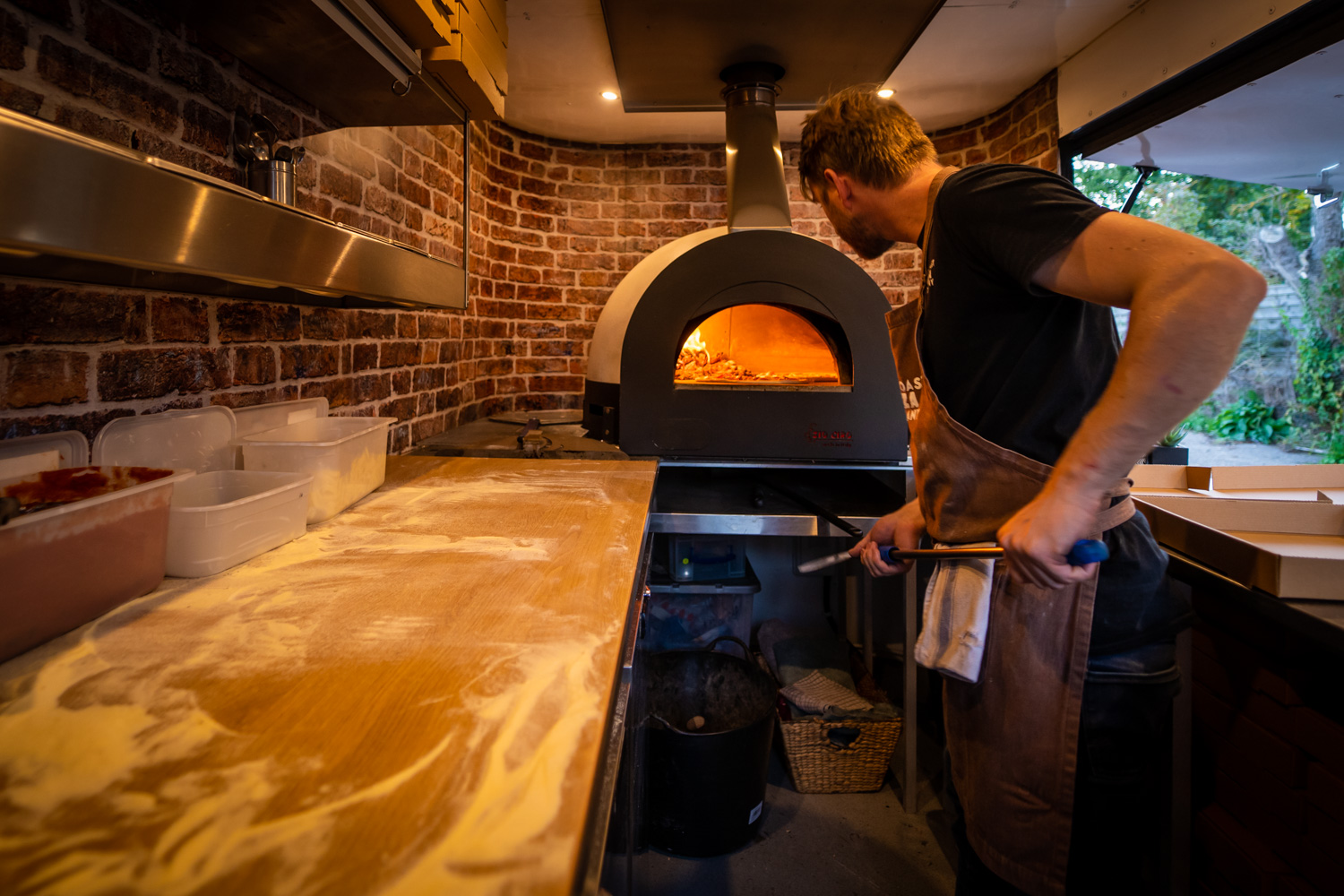 Chris loading a pizza into the glowing wood-fired oven inside the Nag Box