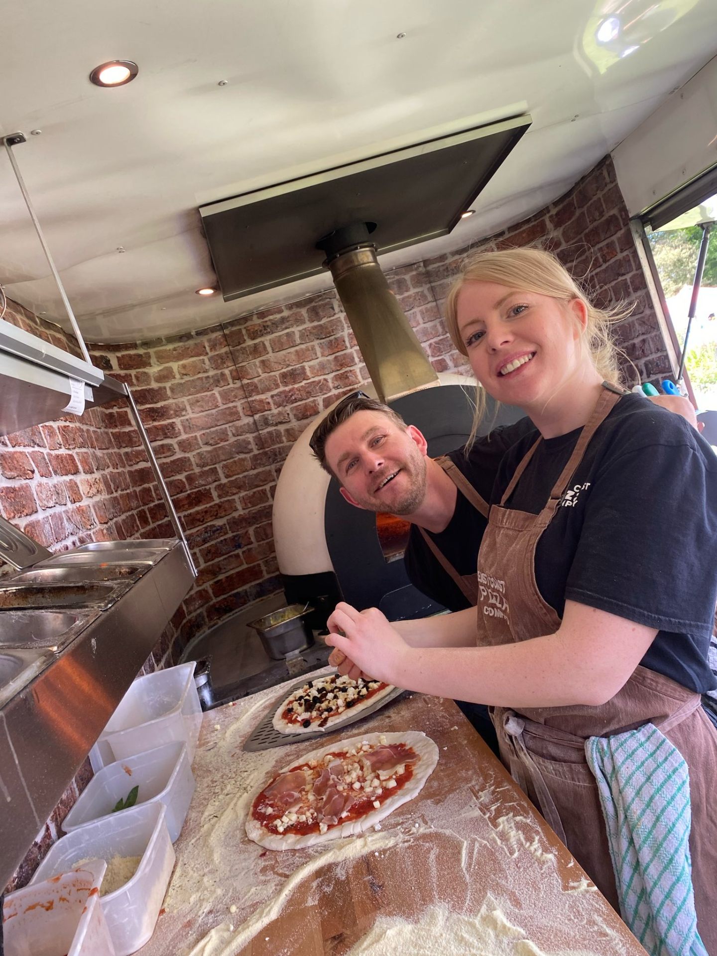 Two of the East Coast Pizza team topping pizzas in front of the wood-fired oven