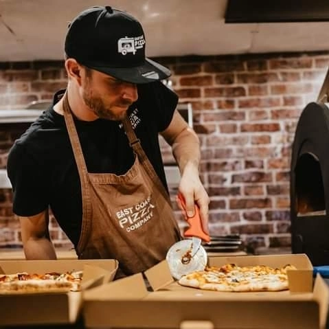 Chris, owner of East Coast Pizza Company, slicing a pizza in his branded apron