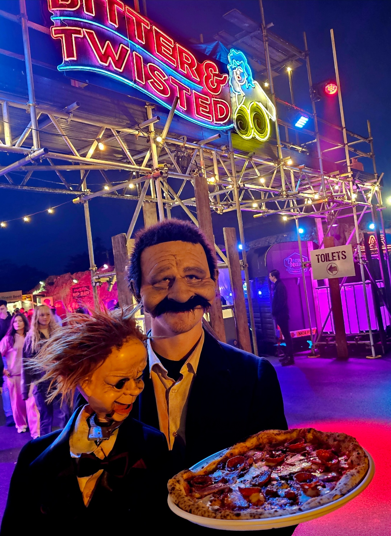 A pepperoni pizza held by a costumed character at PrimEvil scream park under neon lights